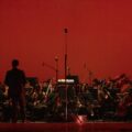 A symphony orchestra performing live on stage under dramatic red lighting.