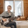 Musician playing electric guitar on a bed in a cozy wooden room with natural light.