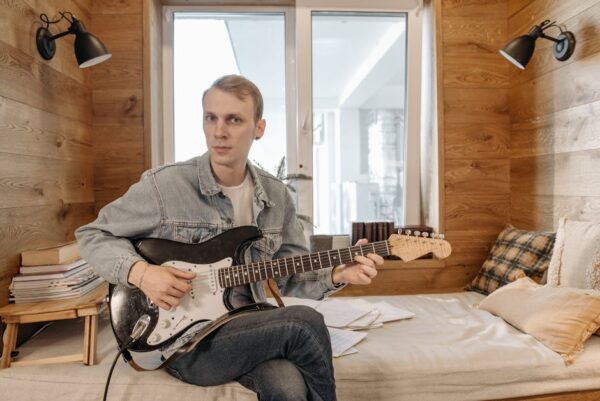 Musician playing electric guitar on a bed in a cozy wooden room with natural light.