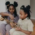 Two children sitting indoors, one teaching the other to play a brass trumpet, creating a warm family moment.
