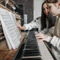 A child learning to play piano with guidance from an adult in a cozy setting, focusing on music sheets.