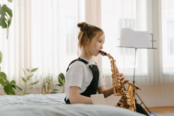 A young girl in overalls playing the saxophone indoors, focusing on music practice.