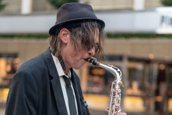 A street musician in a black suit and hat plays saxophone outdoors, creating a lively atmosphere.