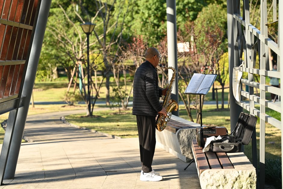 An adult playing saxophone outdoors with sheet music in a serene park environment.