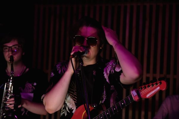 Dynamic image of a live band performance featuring a guitarist, singer, and saxophonist under neon lights.