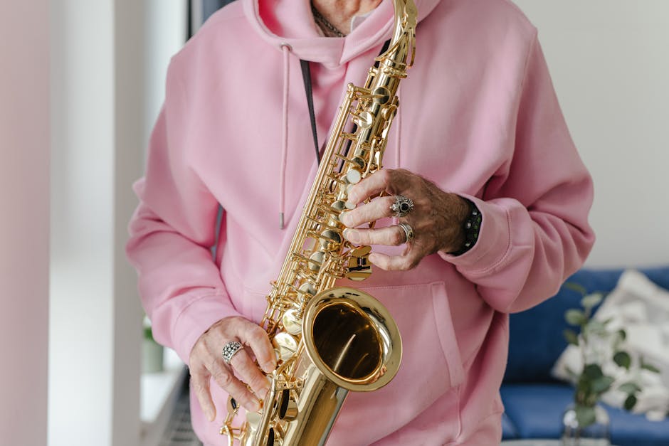A musician plays a saxophone while wearing a pink hoodie indoors.