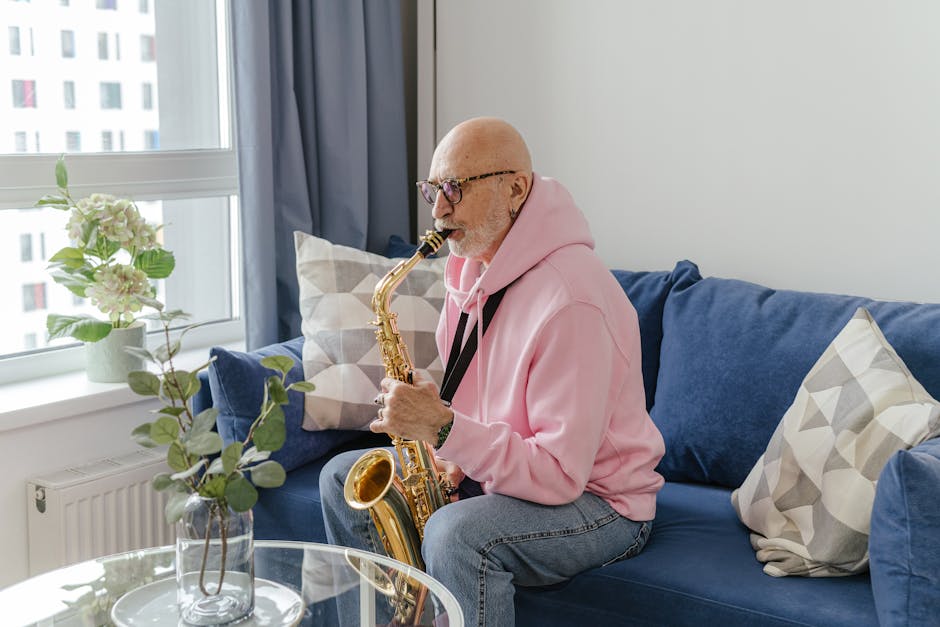 An elderly bald man in a pink sweater plays the saxophone on a couch in a bright living room.