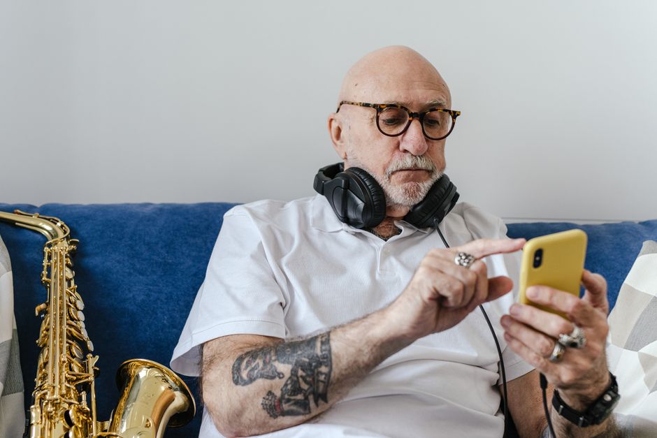 Elderly musician with headphones and saxophone using smartphone indoors.