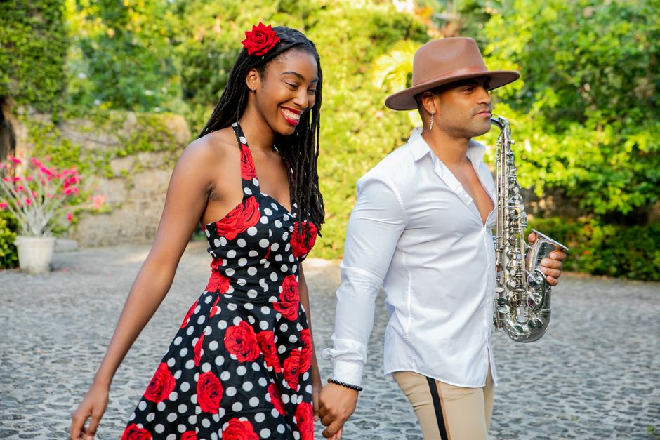 A stylish couple walking hand in hand outdoors; the man carries a saxophone.