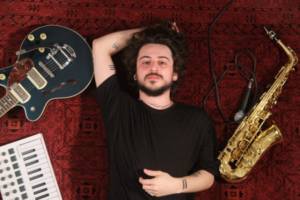 A man lies on a red rug surrounded by a guitar, saxophone, and keyboard, evoking a musical vibe.