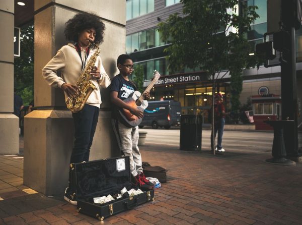 Concentrated African American guy with curly hair in casual clothes standing and playing saxophone with friend guitarist while earning money on city street in evening time