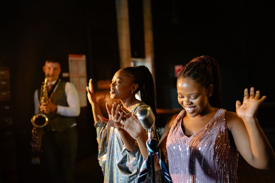 Black women singing energetically with a saxophone player in a lively jazz performance.