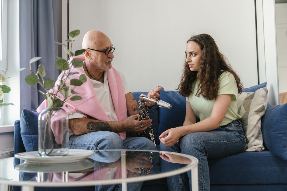 Elderly man and young woman seated on sofa discussing saxophone indoors.
