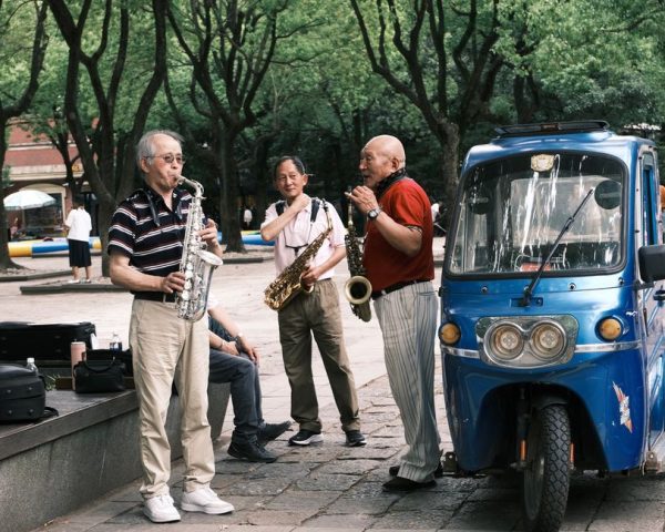 Asian men play saxophones on a city street, performing next to a blue vehicle.