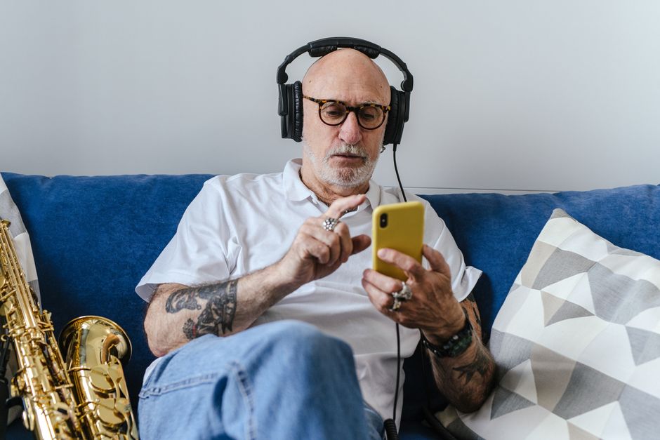Elderly man with tattoos listening to music on headphones while using smartphone, indoor setting.