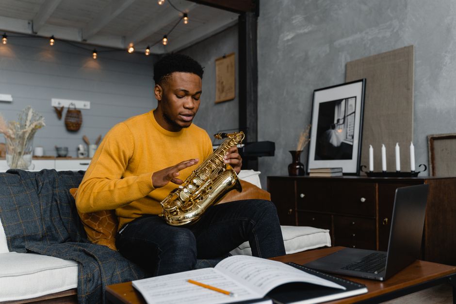 Young man sitting on a couch, practicing saxophone with sheet music in a cozy living room.