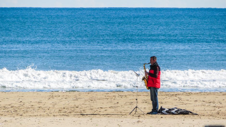 A man plays saxophone on a sandy beach with waves in the background.