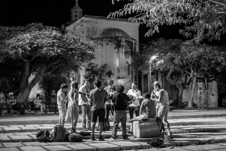 A group of street musicians performing at night in a city square, surrounded by trees and historic architecture.