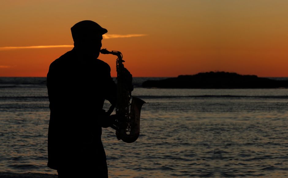 A saxophonist plays by the ocean at sunset, creating a serene silhouette scene.