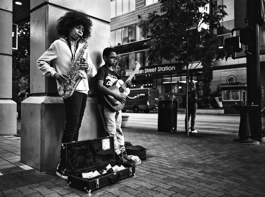 Black and white of African American guy with curly hair playing saxophone and standing near friend playing guitar on city street