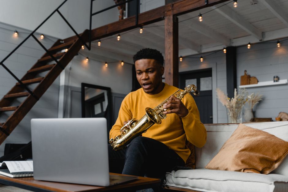 Young man with saxophone learning music online at home, using laptop on couch.