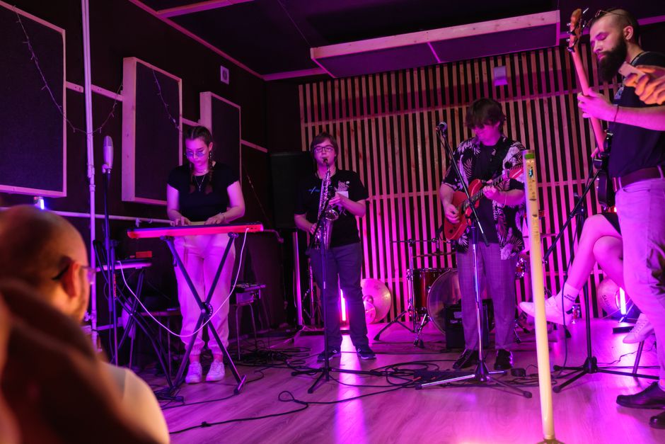Live band performing on a small indoor stage with vibrant lighting, featuring various musicians and instruments.