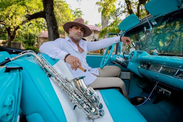 A fashionable man poses with a saxophone in a classic convertible car.