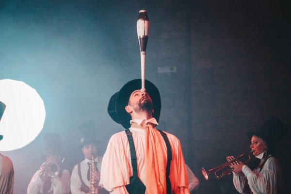 A circus performer balances a juggling club on his chin, surrounded by a band playing music.
