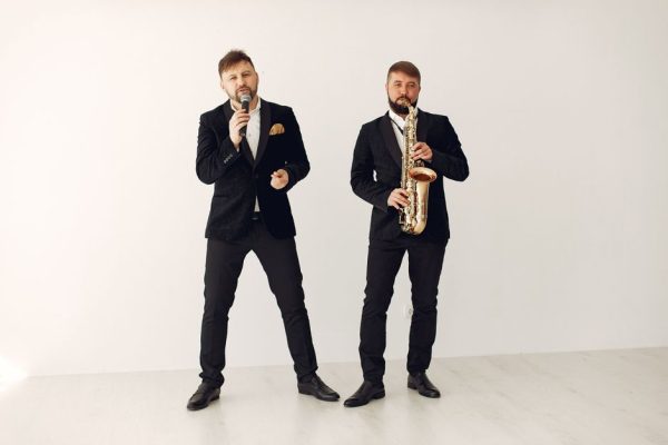Full body adult male musician playing saxophone near inspired male vocalist singing popular song while standing together against white wall during celebration party or live music show
