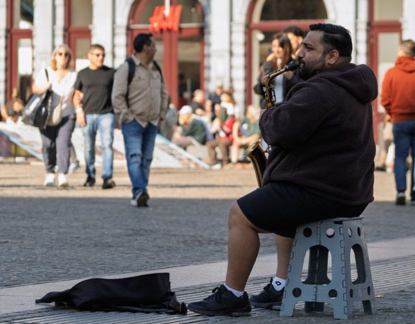 A street musician performs on a saxophone as passersby stroll through a bustling urban square.