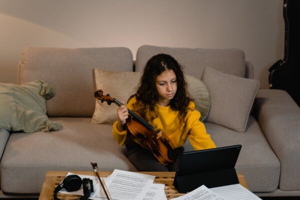 Young girl in yellow sweater learning violin using a tablet on a cozy sofa.