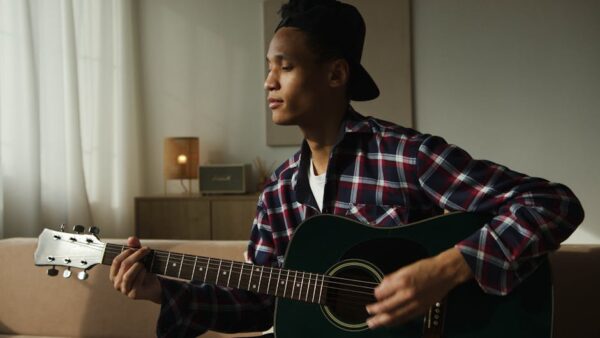 Young man playing an acoustic guitar in a cozy indoor setting, wearing a plaid shirt and cap.