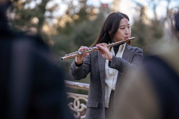 Asian woman in a blazer playing flute outdoors with a blurred background.