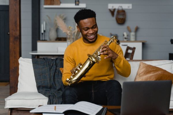 Cheerful man in a yellow sweater playing a saxophone on a couch in a cozy modern living room.