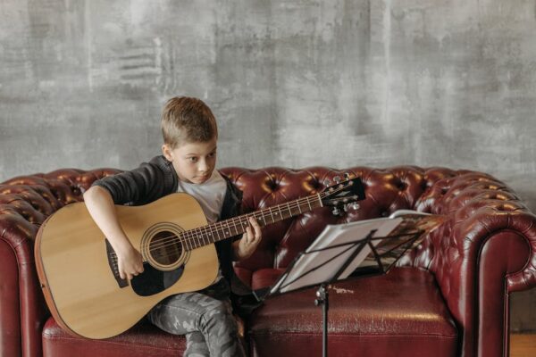 A young boy practicing guitar indoors on a comfy leather sofa, focusing on his music sheet.