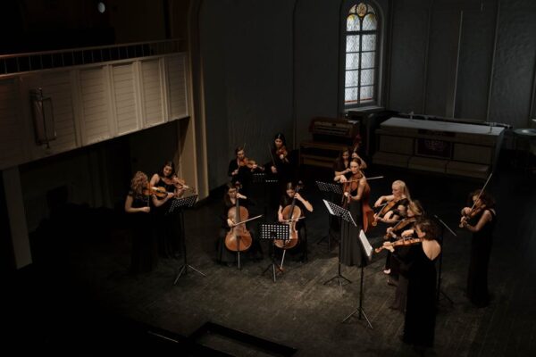 A diverse group of women performs classical music with violins and cellos on an indoor stage.