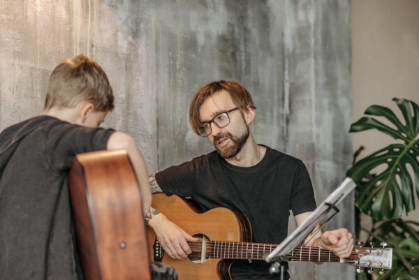A young boy learning guitar from a bearded man indoors, emphasizing musical guidance.
