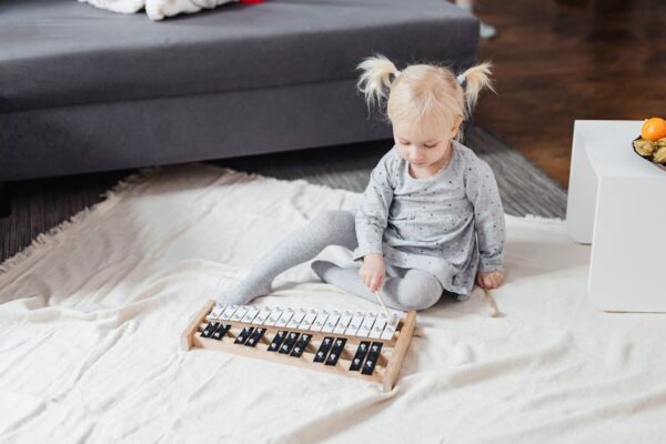 A cute blonde girl sitting on the floor at home playing a xylophone with mallets.