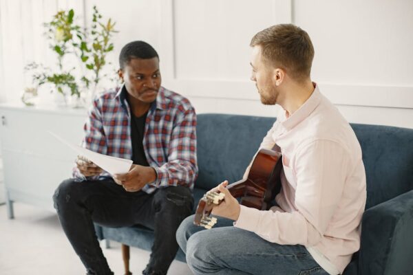 Two men sitting on a couch discussing music with guitar and papers.