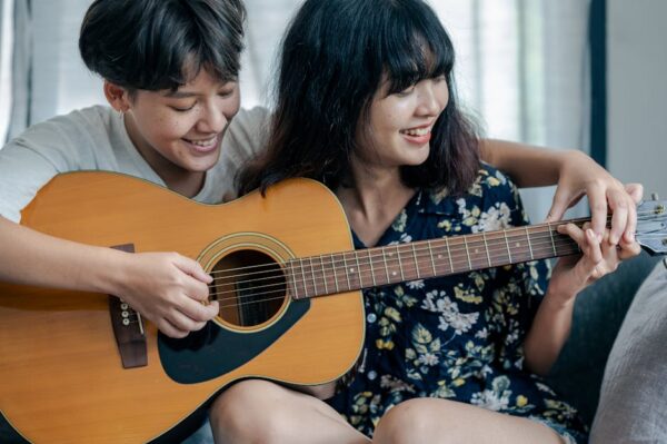 A joyful lesbian couple enjoys playing an acoustic guitar together indoors, showcasing their close relationship and shared passion for music.