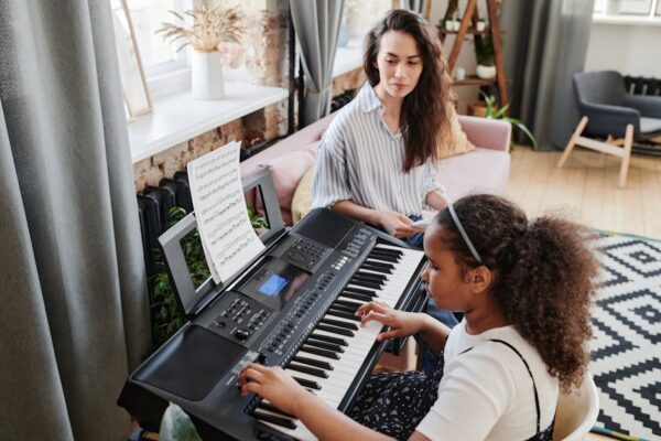 Girl takes piano lessons with attentive teacher observing indoors.