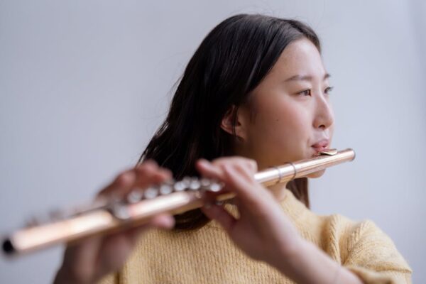 Young Asian lady in warm sweater playing flute in light room with white wall