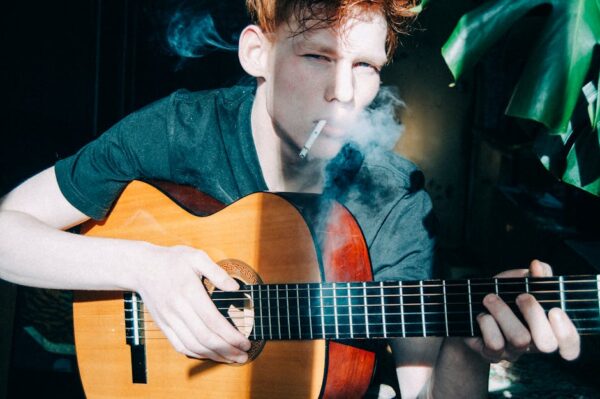 Portrait of a young man playing an acoustic guitar and smoking indoors in soft sunlight.