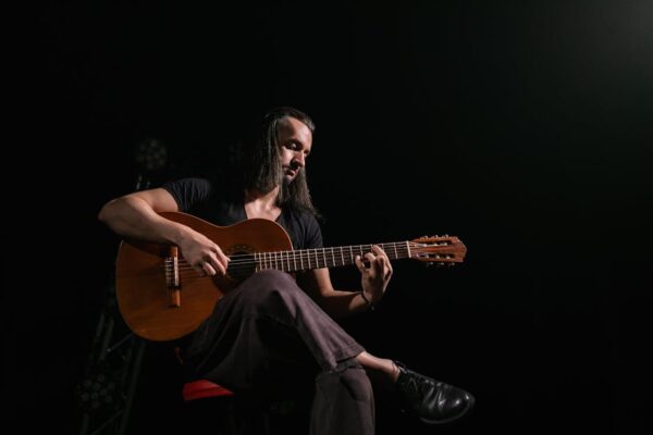 A musician with long hair sitting and playing a guitar in a dark room.