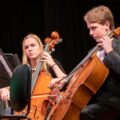 Two young cellists performing in an orchestra with focus and intensity.