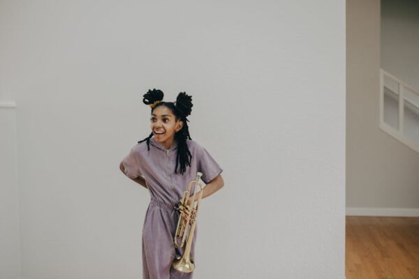 Young woman with afro hair in a purple romper holding a trumpet indoors.