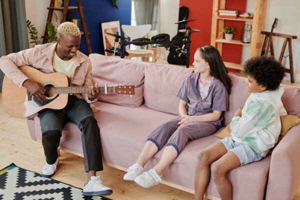 Man teaching guitar to children on a sofa in a cozy living room.