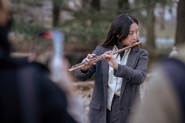 Asian female musician playing flute in an outdoor park setting, surrounded by blurred audience.