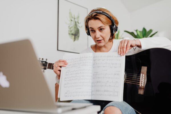 Woman teaching guitar online, presenting sheet music via video call with laptop setup.
