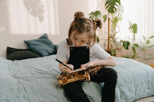 A young girl sits on a bed writing music notes while holding a saxophone.